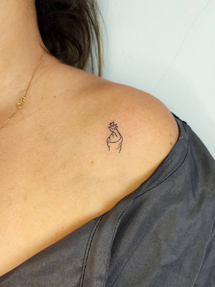 Mulher de pele clara e blusa preta com tatuagem de mão fazendo coração coreano no ombro
