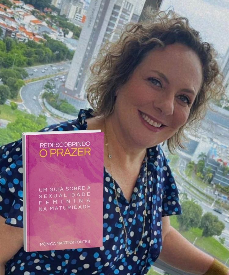 Mônoca Martins Fontes sorridente com cabelos cacheados e vestindo blusa azul de bolinhas posa diante de uma janela com vista urbana ao fundo. Em destaque, a capa rosa do livro "Redescobrindo o Prazer" aparece posicionada na imagem.