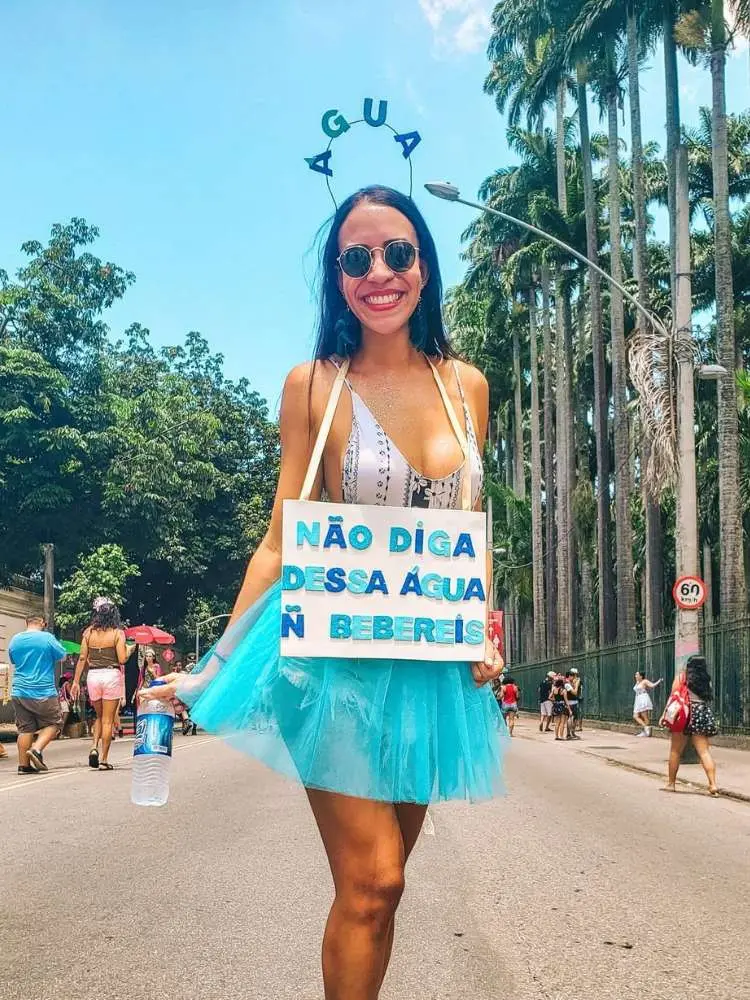 Mulher d pele morena usando body preto e branco, saia azul de tule com tiara escrito "água" e placa escrito "não diga dessa água ñ bebereis"