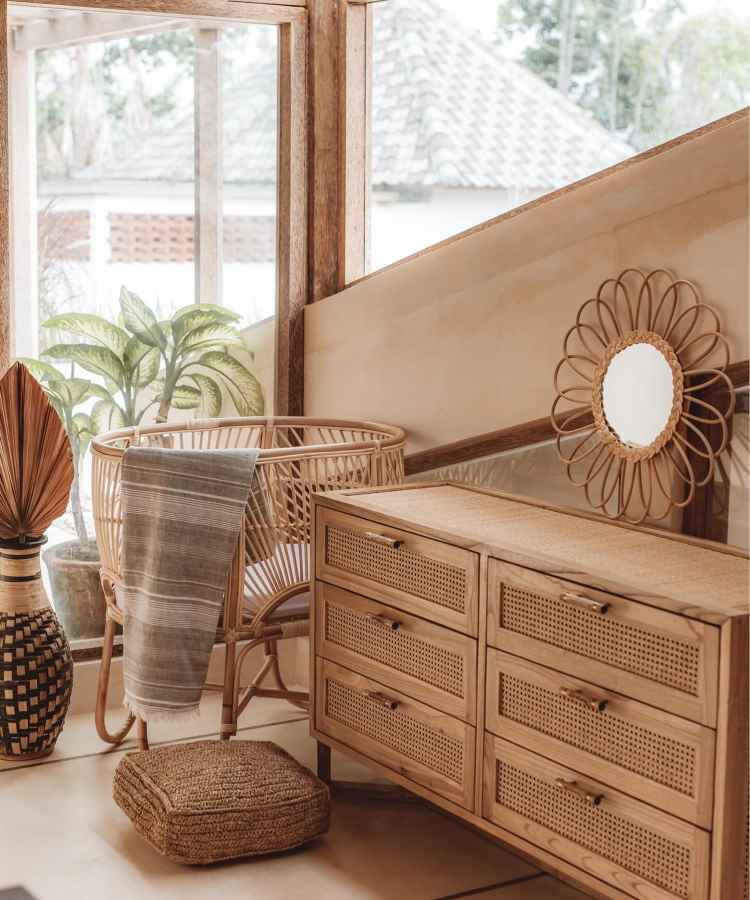 A imagem apresenta um espaço acolhedor com uma cadeira de vime e almofada. Há uma cômoda de madeira com detalhes em palhinha e espelho decorativo. Plantas e um cesto texturizado complementam o ambiente, trazendo um toque natural e rústico. - tendências de decoração para 2025