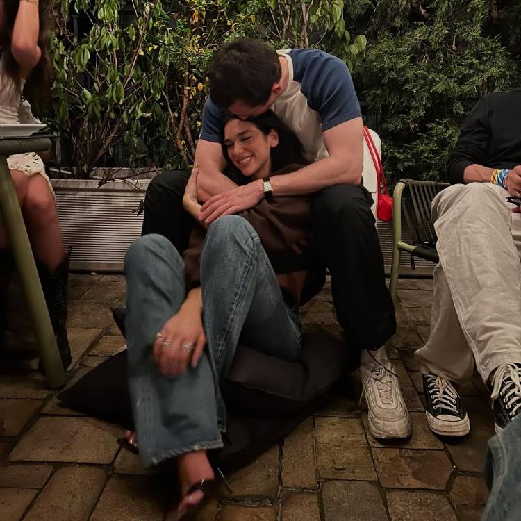 Um casal se abraça em uma área externa, possivelmente um pátio. A mulher está sentada em uma almofada no chão, sorrindo, enquanto o homem a abraça por trás. O casal parece relaxado e feliz.