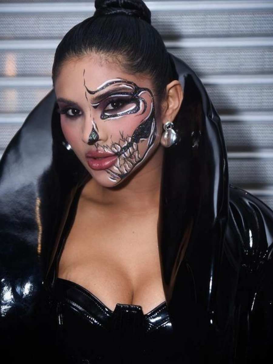 Imagem em fundo com porta de enrolar em aço e Mileide na frente. Ela veste look preto e maquiagem de caveira no rosto.