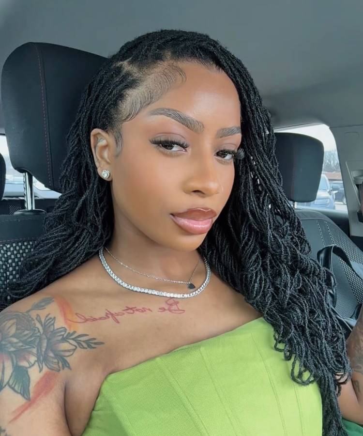 Mulher com cabelos estilo dreadlocks, baby hairs estilizados na testa, maquiagem impecável e colar delicado. Ela usa top verde, exibe tatuagens florais e posa dentro de um carro, transmitindo confiança e elegância em ambiente casual.





