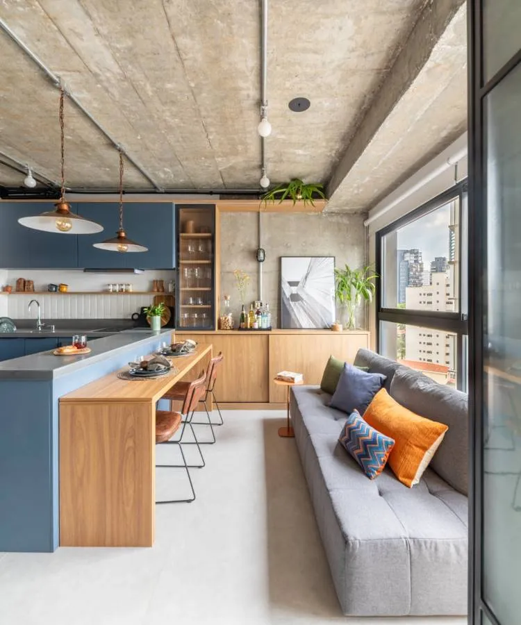 A imagem mostra um interior de apartamento moderno com um layout de plano aberto. O teto é de concreto inacabado, e há duas luminárias pendentes com design simples. À esquerda, há uma área de cozinha com armários azul-escuros e detalhes em madeira. A bancada da cozinha também é de madeira, e há vários bancos com assentos pretos e pernas de madeira ao longo dela. Acima da bancada, prateleiras abertas seguram vários itens de cozinha.

Ao lado da cozinha, há uma área de jantar com uma mesa de madeira e cadeiras com estofado laranja e estruturas pretas. Uma grande janela ocupa quase toda a parede direita, proporcionando muita luz natural e uma vista dos prédios do lado de fora.

Em primeiro plano, há uma área de estar com um sofá cinza adornado com almofadas de padrões geométricos em tons de azul e laranja. Em frente ao sofá, há um tapete em cores suaves e, à direita, uma pequena mesa lateral com itens decorativos.