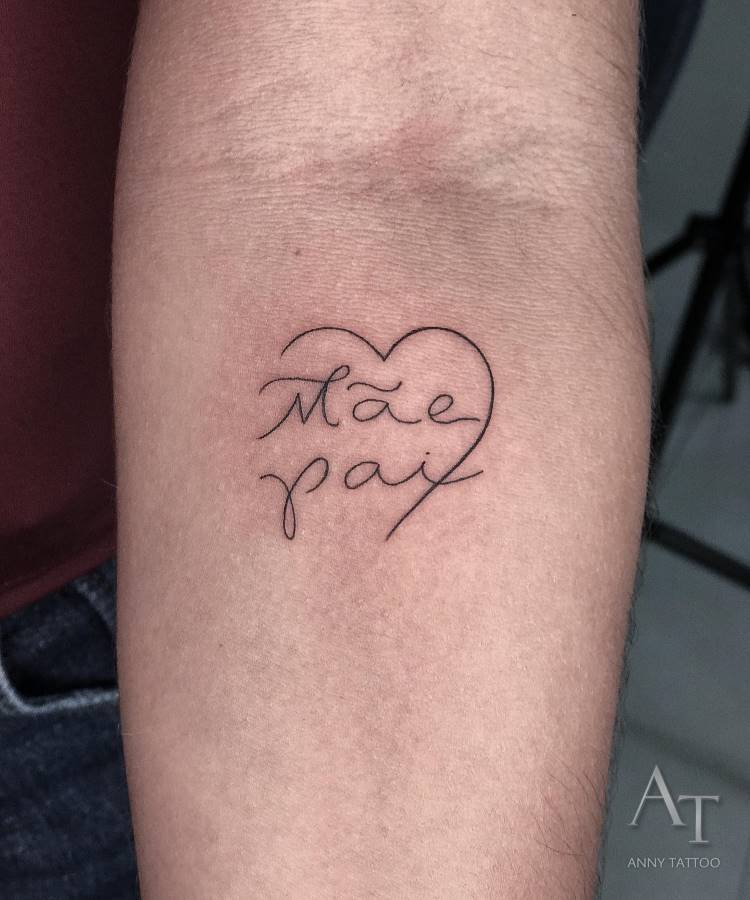 A imagem exibe uma tatuagem no antebraço de uma pessoa, consistindo no contorno de um coração com as palavras "Mãe" e "Pai" escritas em caligrafia cursiva dentro dele. A tatuagem é em preto, e a pele é clara. No canto inferior direito, há uma assinatura digital "AT ANNY TATTOO".







