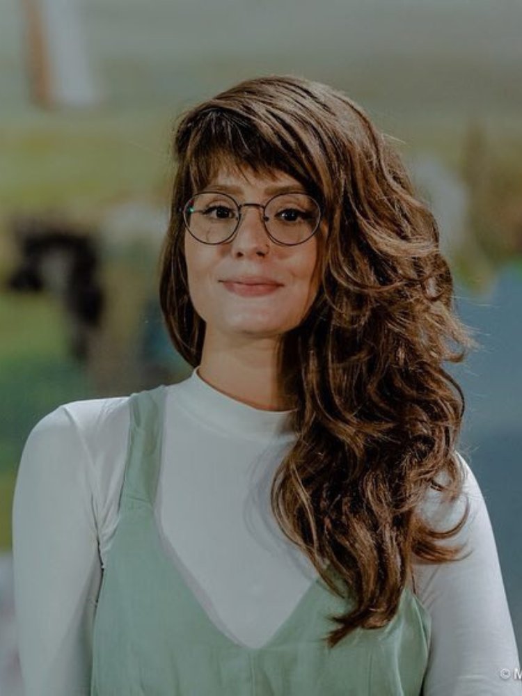 Thais Dutra Sá em 2019, com cabelo longo de lado, óculos redondo, blusa branca e salopete verde