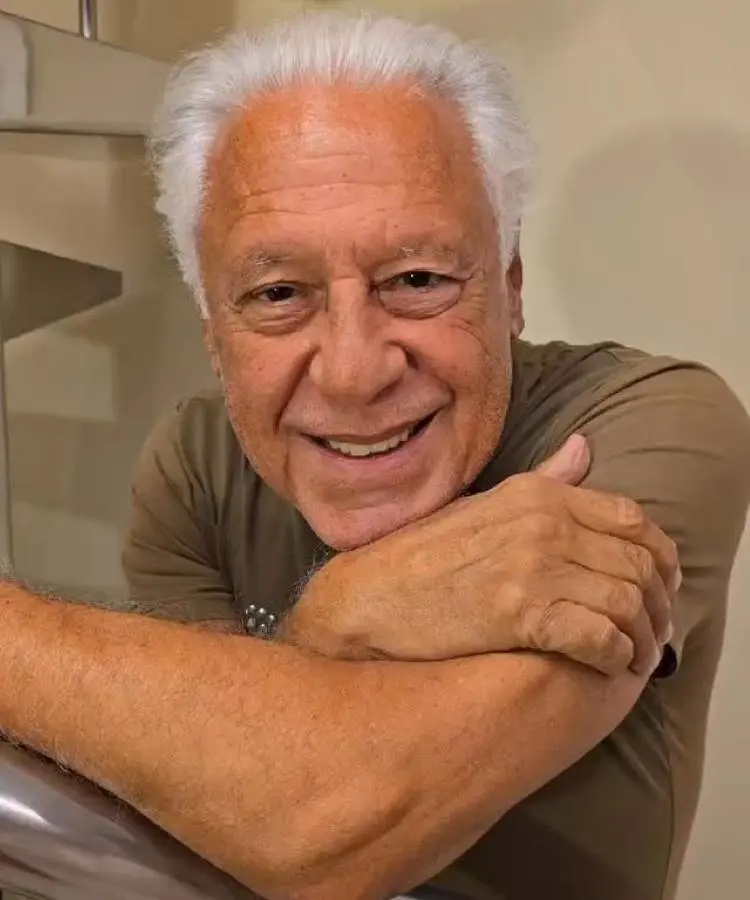Homem de cabelos brancos curtos e sorriso acolhedor, com os braços cruzados apoiados em uma superfície metálica. Usa camiseta bege em ambiente interno com decoração discreta ao fundo. Ícone do elenco de "Vale Tudo", hoje admirado por sua carreira.