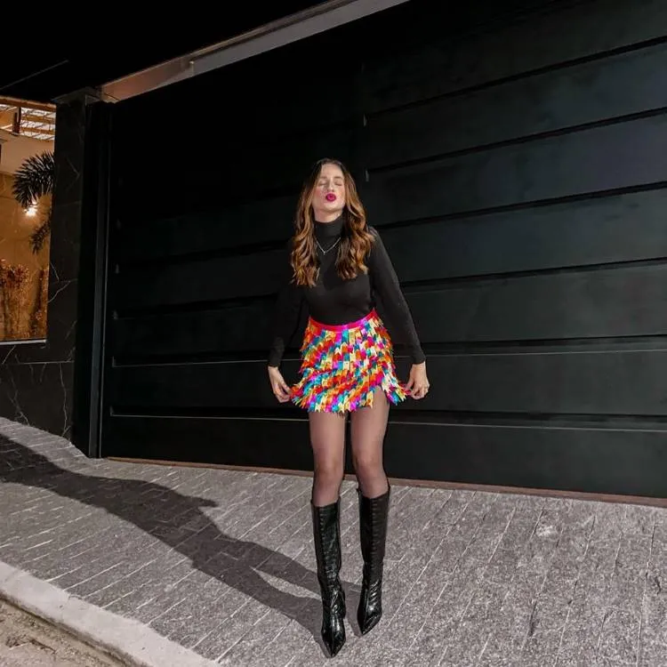 Mulher de cabelos ondulados castanhos e caramelo, com roupa preta e saia multicolorida, posa em frente a porta de garagem moderna. Usa botas pretas brilhantes e meia-calça. Ao lado, janela com iluminação quente e decoração. Atmosfera noturna urbana.
