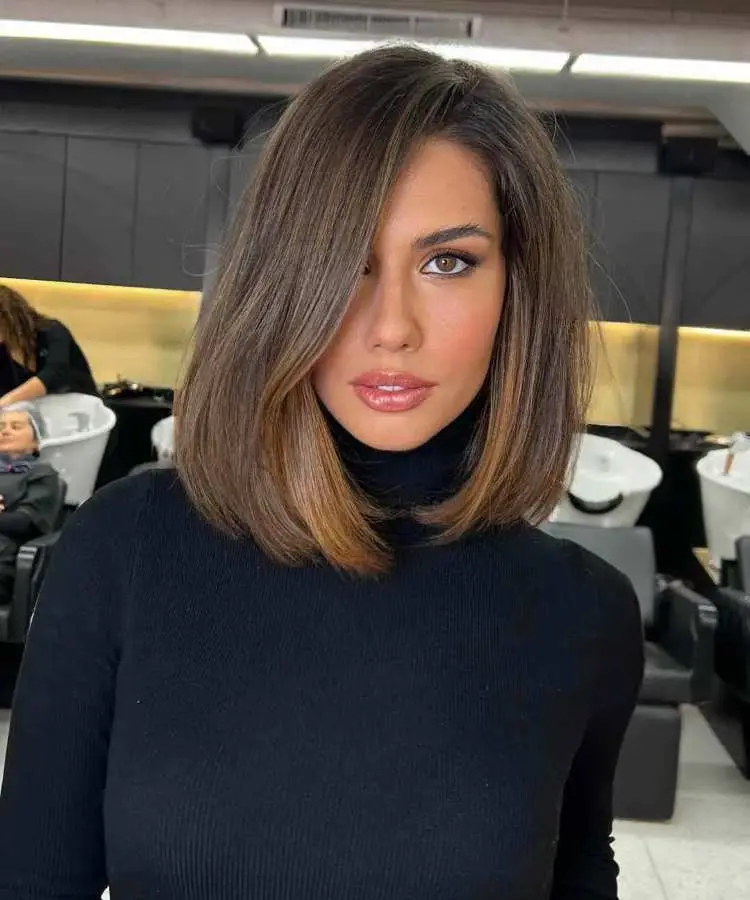 Mulher com cabelo médio castanho, corte reto e polido, com leve curvatura nas pontas e mechas iluminadas. Divisão lateral destaca o rosto com maquiagem suave. Ela usa blusa preta de gola alta. Fundo interno em salão de beleza.