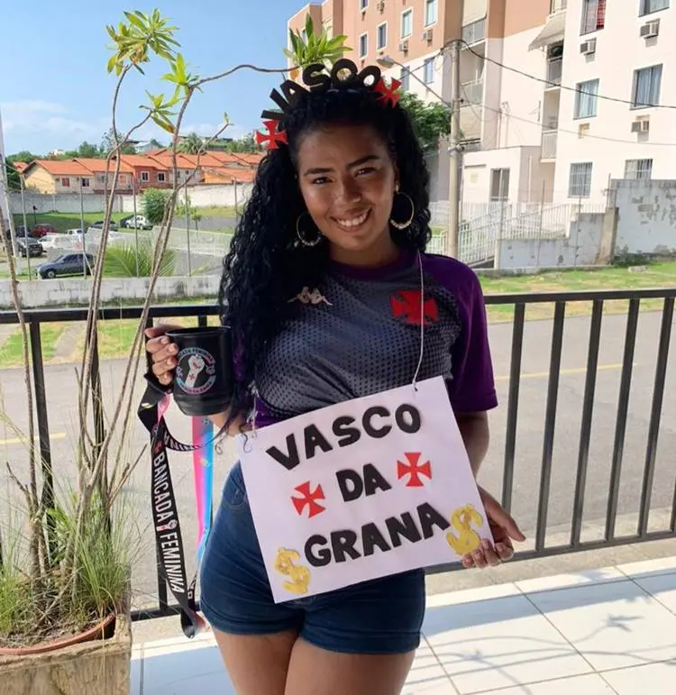 Mulher de pele negra com camisa do vasco, shorts jeans, tiara do Vasco e plaquinha "Vasco da Grana"