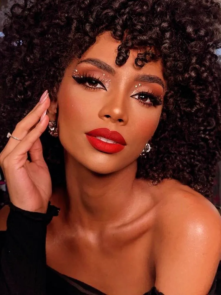 Mulher de pele negra usando maquiagem levemente vermelha nos olhos, com alguns brilhos colado e batom vermelho. 