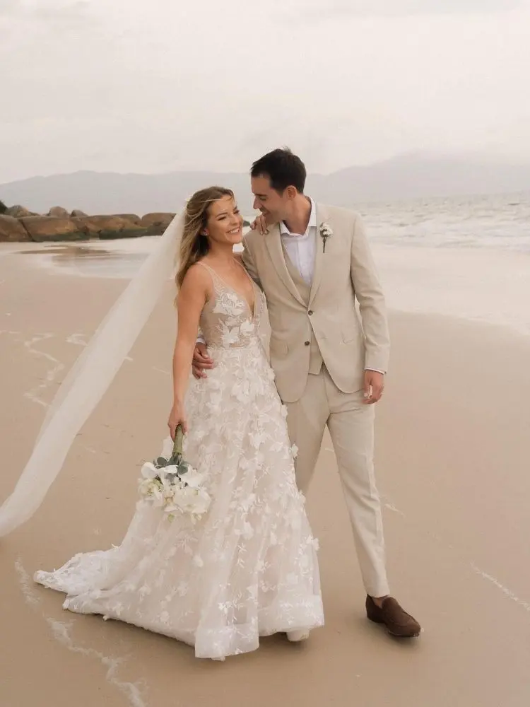 Mulher de pele clara usando véu, vestido de noiva de tule com bordados 3D e homem de terno bege, comemorando casamento na areia da praia  