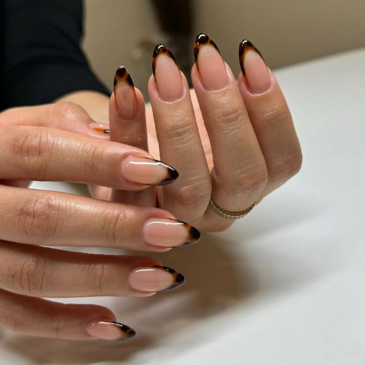 A imagem mostra unhas alongadas e elegantes, decoradas com pontas em estilo tartaruga, em tons de marrom e preto, sobre base nude. A mão exibe um anel dourado delicado. O fundo neutro destaca o design moderno e refinado.