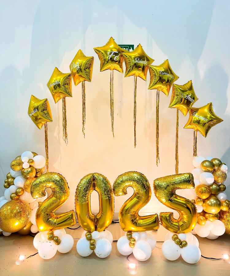 A imagem mostra uma decoração de Ano Novo com balões dourados formando "2025". Estrelas douradas com palavras como "Família", "Paz" e "Saúde" estão no topo. Balões brancos e dourados decoram a base, criando um cenário festivo.


