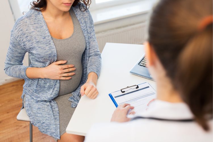 Mulher de pele clara usando vestido cinza e jaqueta jeans, sentada em cadeira em frente à médica de pele clara, que segura uma prancheta