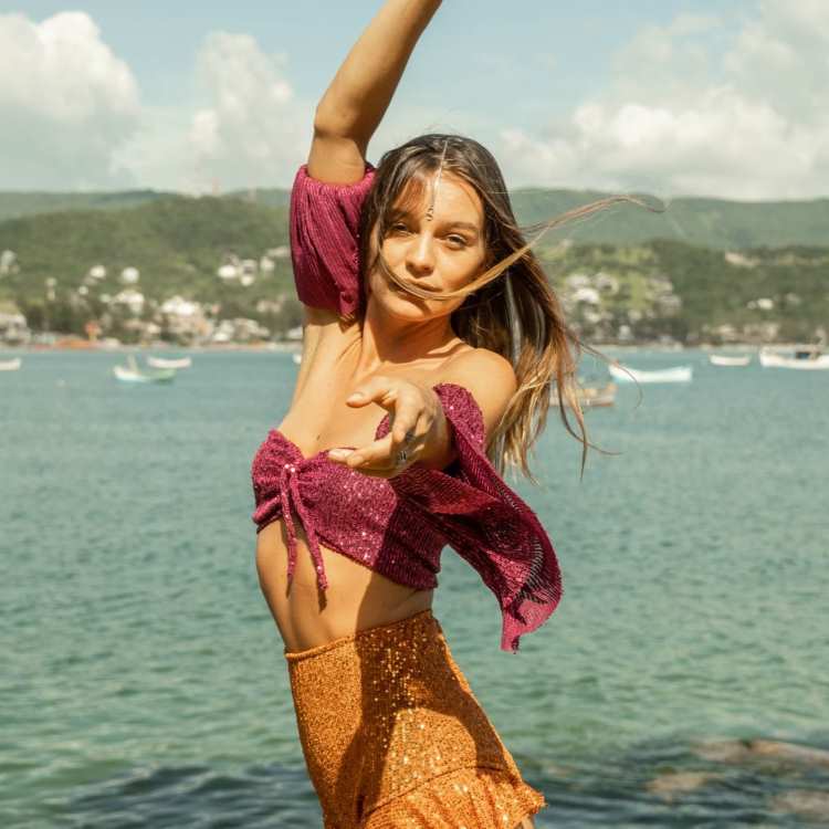 Mulher jovem dança em frente ao mar, usando blusa rosa brilhante e saia dourada com lantejoulas. Sua expressão transmite alegria e leveza, com fundo de barcos, montanhas e céu azul. Cabelos soltos esvoaçam ao vento.