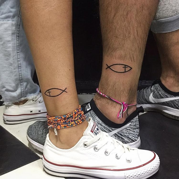 A imagem exibe duas pessoas com uma tatuagem religiosa no tornozelo, representando o símbolo do peixe, conhecido como Ichthys. Este símbolo é um antigo emblema cristão, simbolizando a fé. As tatuagens são simples e elegantes, destacando-se sobre a pele. Ambas as pessoas usam tênis e pulseiras coloridas, criando um visual descontraído.


