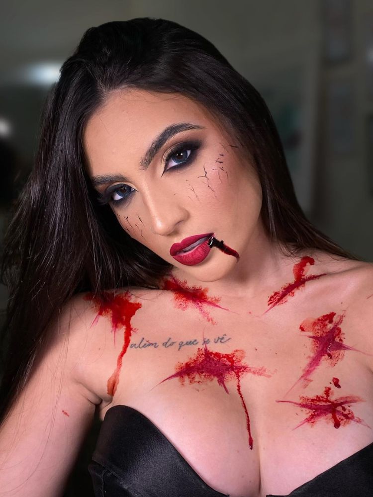 Mulher de pele clara com sangue fake escorrendo da boca e vários machucados com esse sangue no colo
