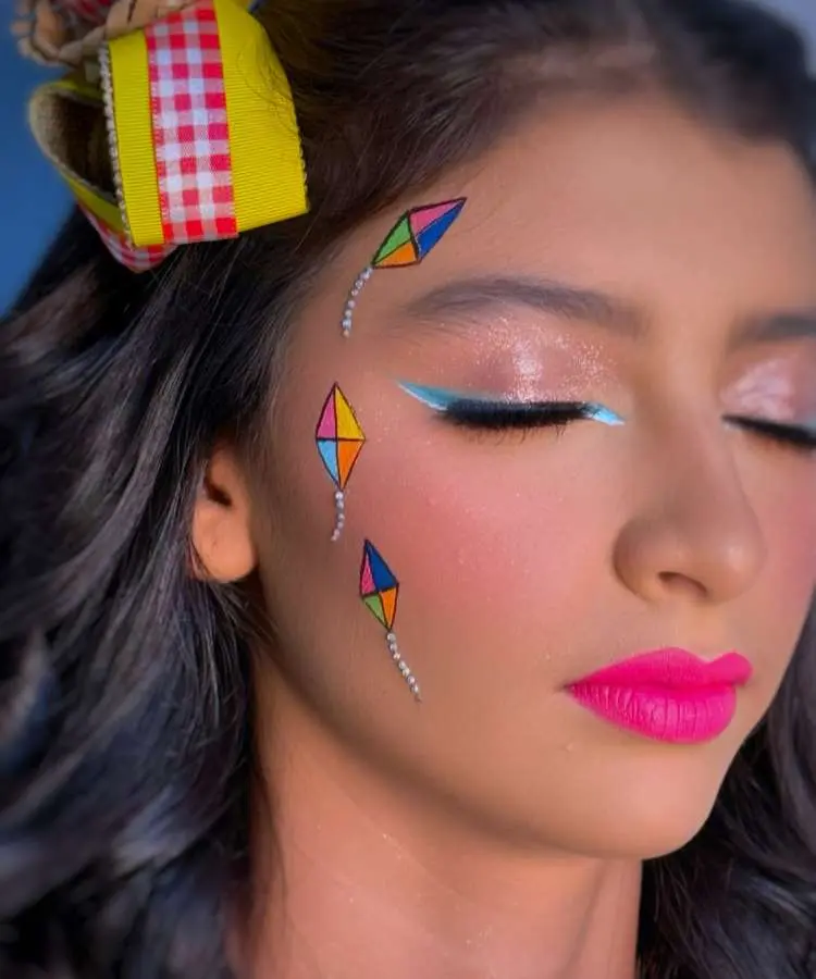 Uma mulher jovem com uma maquiagem criativa de Festa Junina, apresentando delineado gráfico azul-claro nos olhos e pipas coloridas desenhadas na têmpora. Seus lábios têm um batom rosa intenso, e ela usa um laço xadrez amarelo no cabelo.