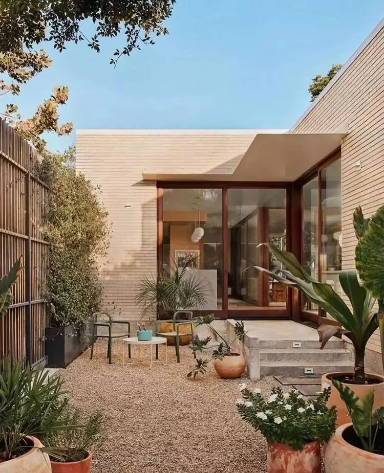 Imagem de fachada de casa simples por dentro. Jardim vertical, flores e plantas em vasos espalhados por chão de pedrinhas. Uma escada de 3 degraus leva para a porta de entrada com vidros grandes