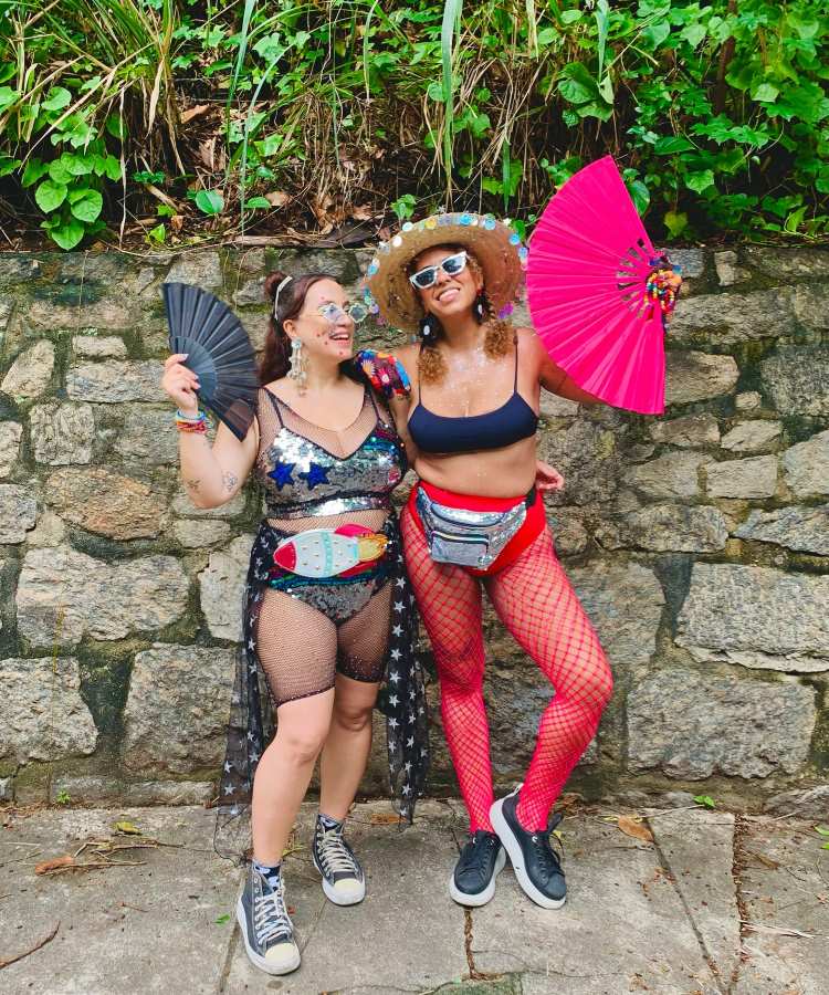 Duas mulheres sorridentes posam juntas em frente a um muro de pedras com vegetação ao fundo. Vestem roupas carnavalescas: uma com vestido de estrelas, outra com top, meia arrastão e chapéu decorado. Ambas seguram leques coloridos, transmitindo alegria.