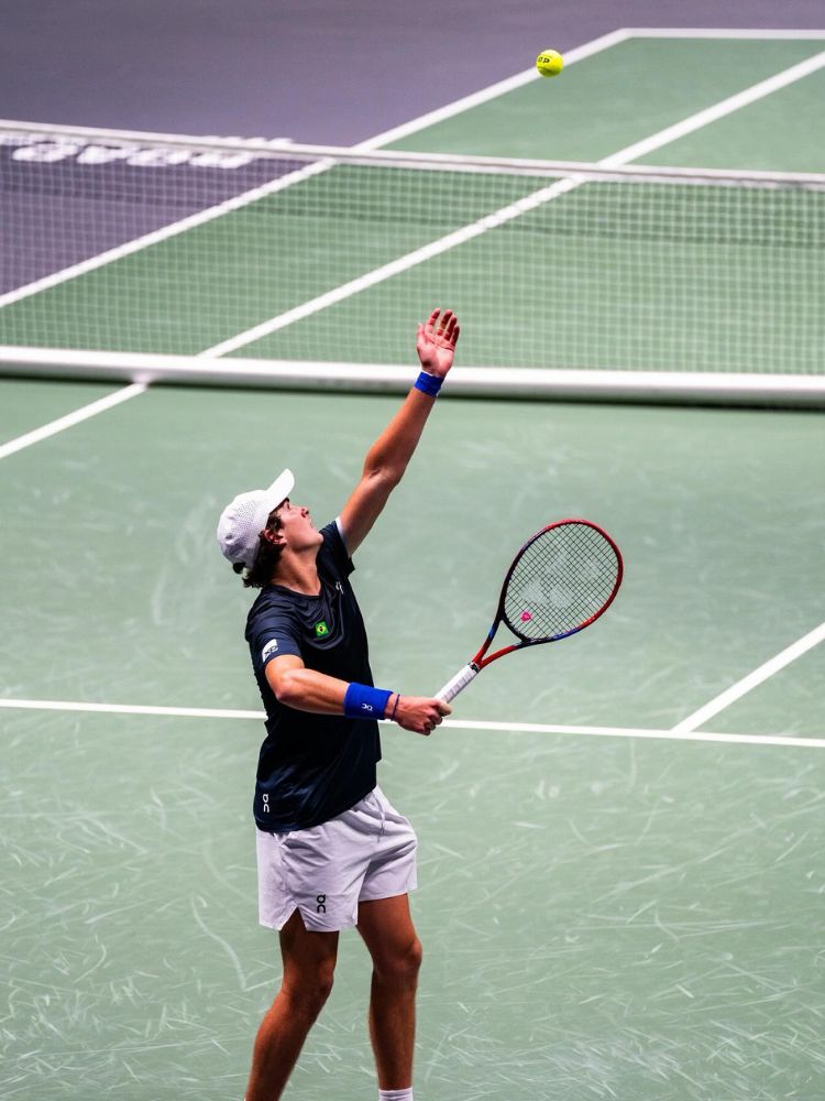 João Fonseca de blusa preta e boné e bermuda brancas sacando com raquete Yonex VCORE 98