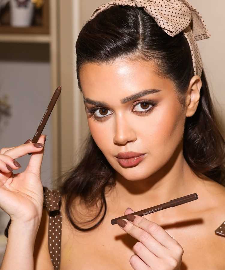 Franciny Ehlke com maquiagem impecável segura dois lápis para olhos marrons com a inscrição "Choco Liner". Ela veste roupa marrom de bolinhas e usa laço combinando nos cabelos. Ao fundo, prateleira desfocada com elementos decorativos.
