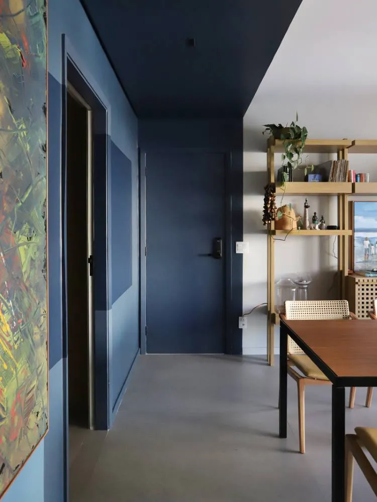 Esta imagem mostra um corredor com paredes e um teto colorido em tons de azul escuro. Uma porta azul discreta se destaca. À direita, um ambiente com prateleiras de madeira, objetos decorativos e uma mesa de jantar sugere uma sala de estar. Uma obra de arte vibrante adiciona contraste.


