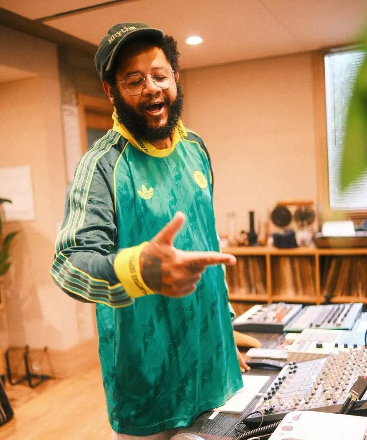 Um homem em estúdio de gravação, barba cheia, óculos e boné verde com "Rhythm". Veste camisa verde da Adidas. Gesticula animadamente. Ao fundo, prateleira com discos de vinil, console analógico e luz suave, criando um ambiente de produção musical acolhedor.
