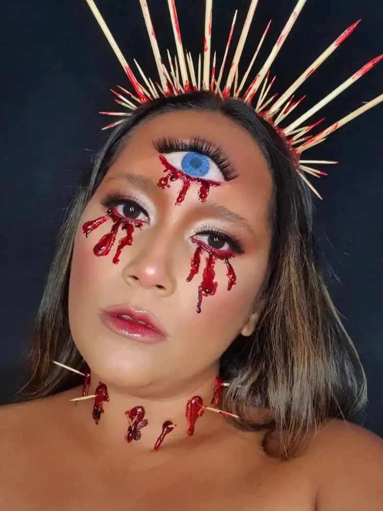 Mulher de pele clara com sangue falso escorrendo dos olhos, desenho de tolho na testa também escorrendo, palitos no pescoço simulando perfurações de estacas e tiara de palitos