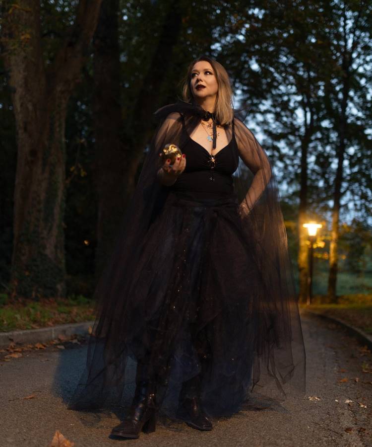 A imagem mostra uma mulher em um cenário noturno, cercada por árvores. Ela veste um vestido preto longo e esvoaçante, com um colar chamativo. Segura uma maçã dourada na mão direita, enquanto olha para cima, com expressão contemplativa. A iluminação suave de um poste ao fundo cria um ambiente misterioso e encantador.


