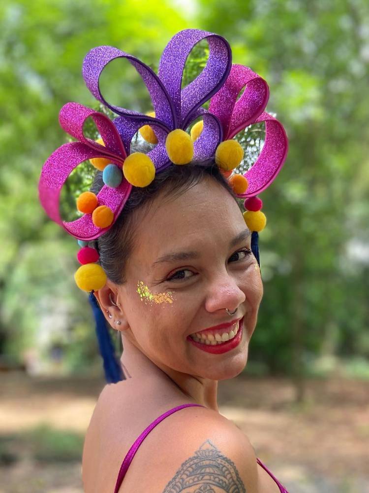 Mulher de pele clara e cabelo castanho preso com tiara de pompom colorido com detalhes em EVA rosa e roxo.