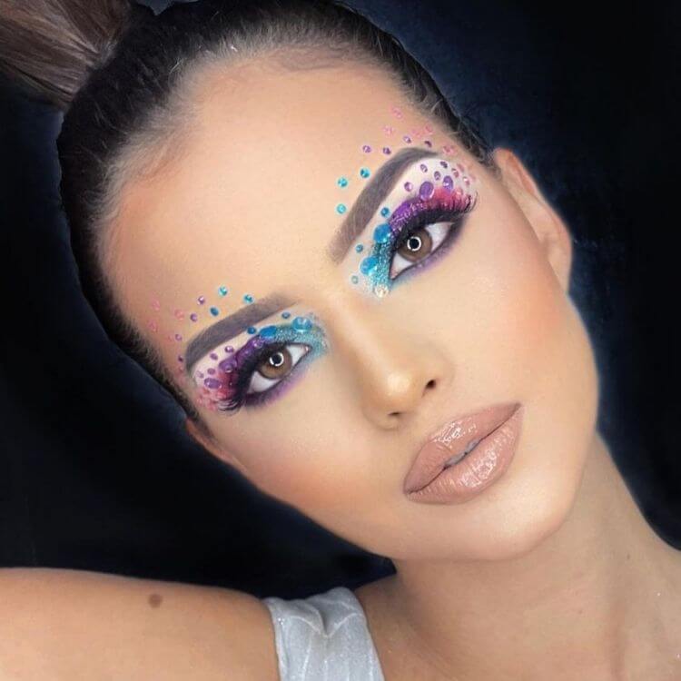 Mulher de pele clara com maquiagem em azul, roxo e rosa, além de strass de diferentes tamanhos acima e abaixo das sobrancelhas e nas pálpebras