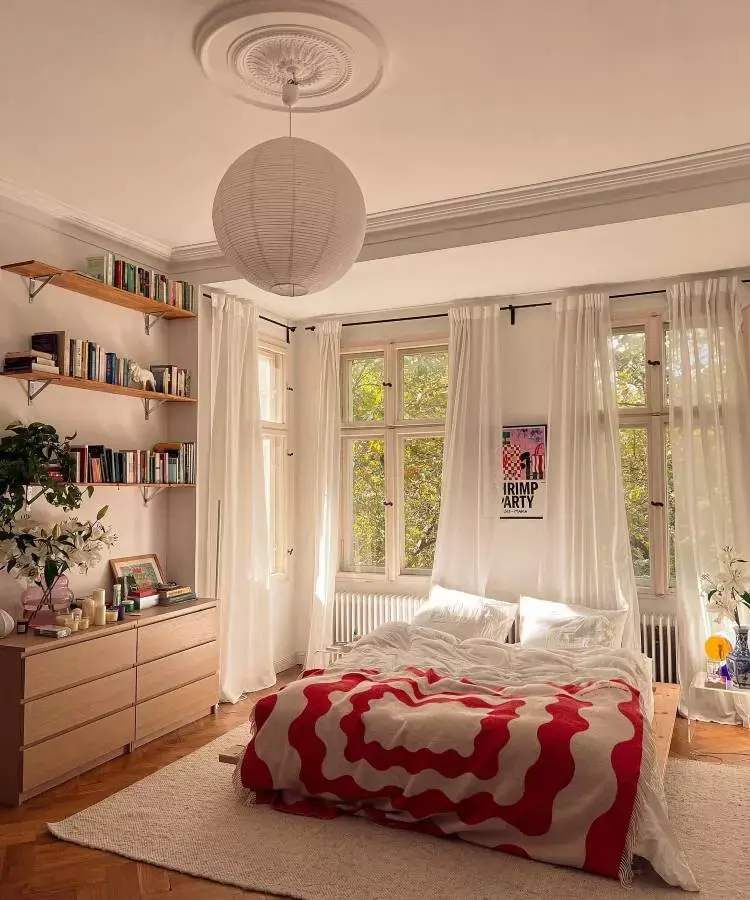Quarto de casal aconchegante, com cortinas brancas, prateleiras de livros e cômoda com velas e objetos