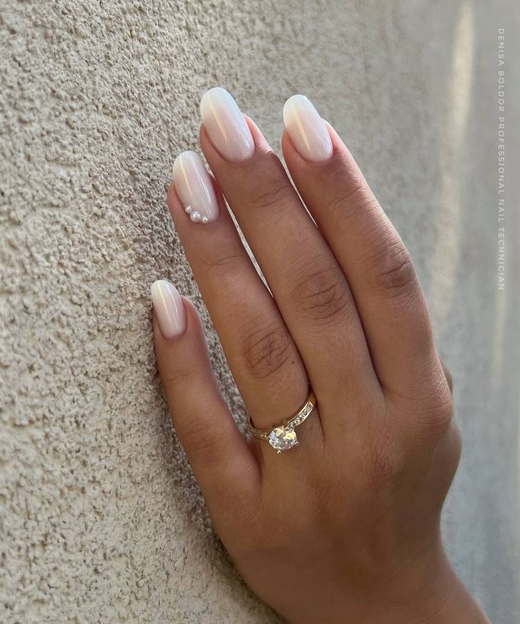 Mão feminina com unhas ovais decoradas com esmalte branco perolado e detalhes em pérolas delicadas no dedo anelar. Anel dourado com pedra central brilhante complementa o visual. Fundo de textura rugosa realça o design elegante e moderno.






