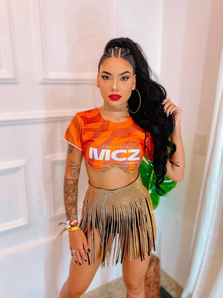 Mulher de pele clara e cabelo escuro usando camiseta de abadá customizado na cor laranja, saia de brilho e hot pant preta.