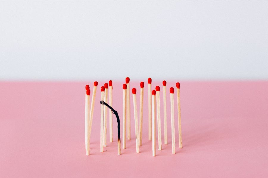 Fundo rosa com vários palitos de fósforo em pé. Um deles está queimado, simbolizando o que é burnout