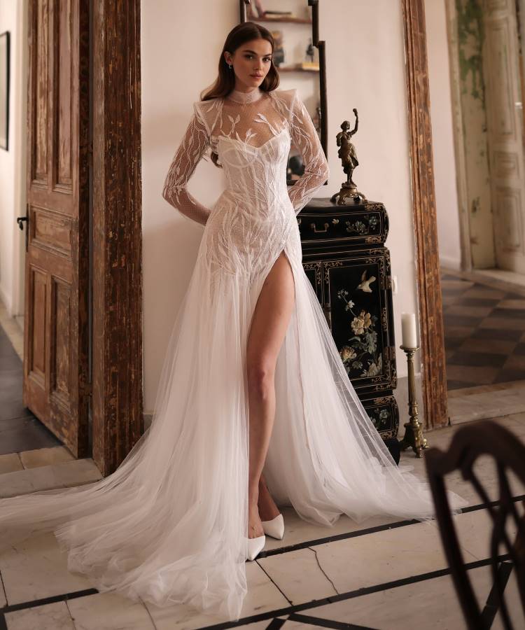 Noiva em vestido branco com tule transparente, bordados florais delicados, mangas longas e fenda alta, representando as tendências em vestidos de noiva 2025. O cenário clássico com móveis elegantes e portas de madeira realça o visual sofisticado.