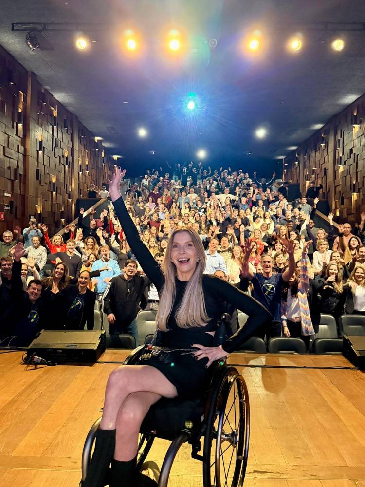 Andrea Schwarz de look all black em palco,, sorrindo e com as mãos para cima, e plateia ao fundo