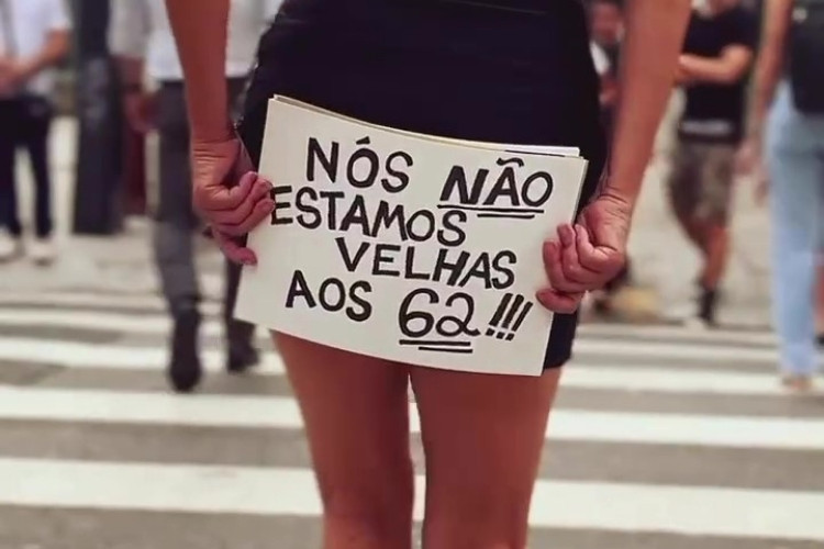Mulher segura um cartaz escrito “Nós não estamos velhas aos 62!!!” enquanto caminha por uma faixa de pedestres na Avenida Paulista, destacando um protesto contra o etarismo. Ela veste saia curta e atrai a atenção de pedestres ao fundo.






