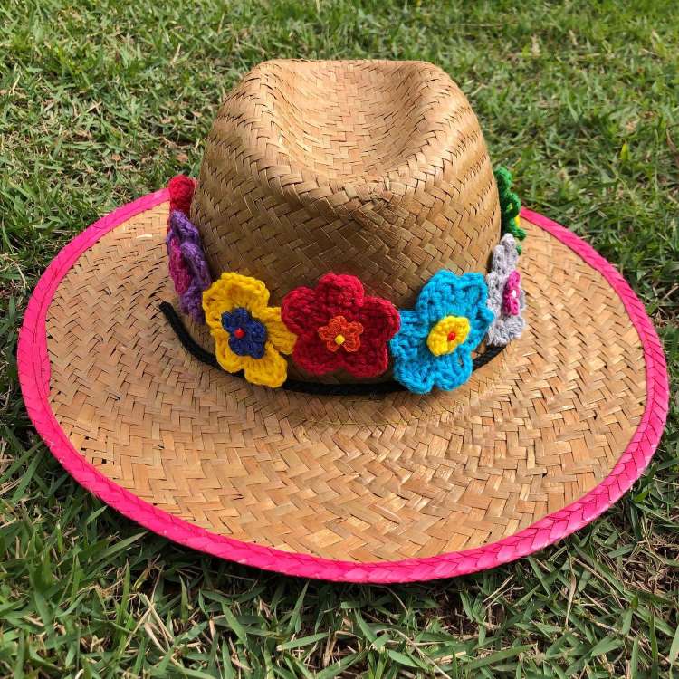 Um chapéu de palha estilo fedora está sobre a grama. Possui uma borda rosa choque e uma faixa preta decorada com flores de crochê coloridas em tons de amarelo, vermelho, azul e cinza.
