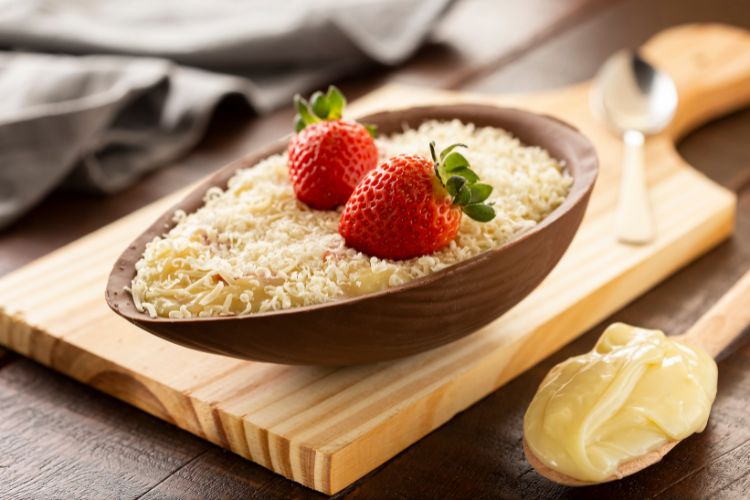 Metade de um ovo de chocolate recheado com creme branco e coberto por raspas de chocolate branco, decorado com dois morangos frescos. O ovo está sobre uma tábua de madeira, com uma colher de creme ao lado.
