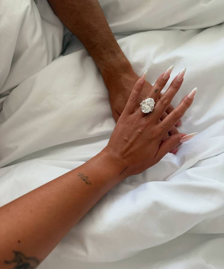 Mãos de um casal se entrelaçam sobre um lençol branco. A mão feminina em primeiro plano exibe uma unha francesa longa e um anel de noivado enorme e brilhante com uma pedra oval. Famosas que estão noivas