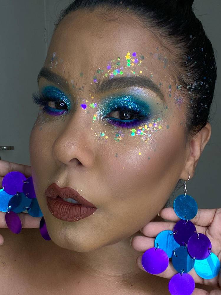 Mulher de pele clara e cabelo preso com sombra de glitter azul, brilho ao redor dos olhos e brinco de círculos azuis. 
