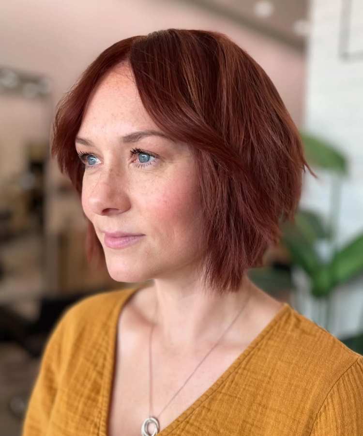 Mulher de cabelo curto estilo bob ondulado em tom de vermelho-acobreado, com franja repartida ao meio, pele clara com sardas, olhos azuis destacados, maquiagem leve, usando blusa amarelo-mostarda e colar minimalista, posando em ambiente iluminado.
