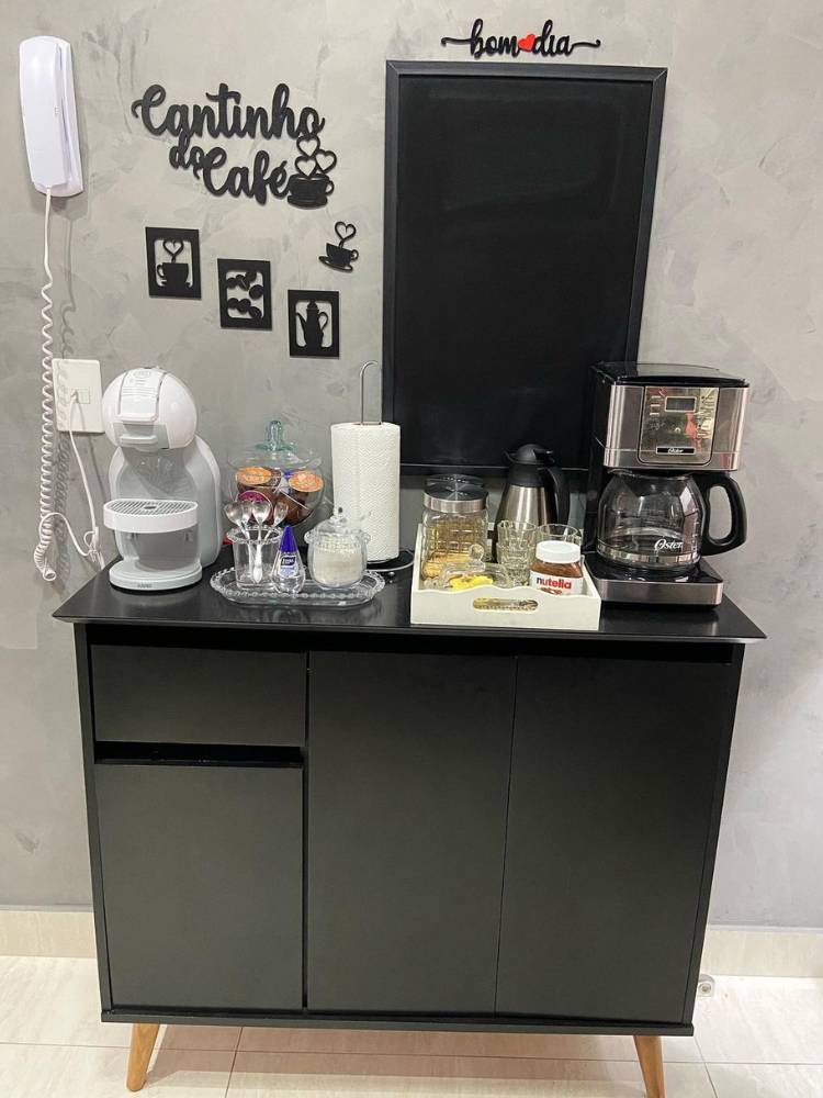 Cantinho do café com quadros na parede, armário preto, cafeteira preta e outra branca, e diversos itens de café