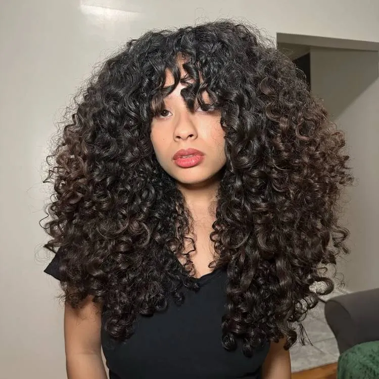 Uma mulher com longos cabelos cacheados escuros e franja olha para a frente, com os lábios ligeiramente abertos. Ela veste uma blusa escura e posa para a câmera.