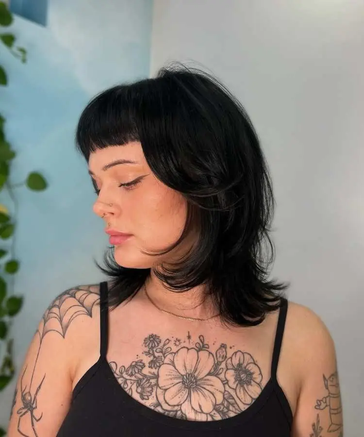 Mulher com cabelo médio preto, corte em camadas com pontas modeladas e franja curta reta. Ela veste regata preta, exibindo tatuagens florais no colo e teia de aranha no ombro. Fundo iluminado com plantas e parede clara.