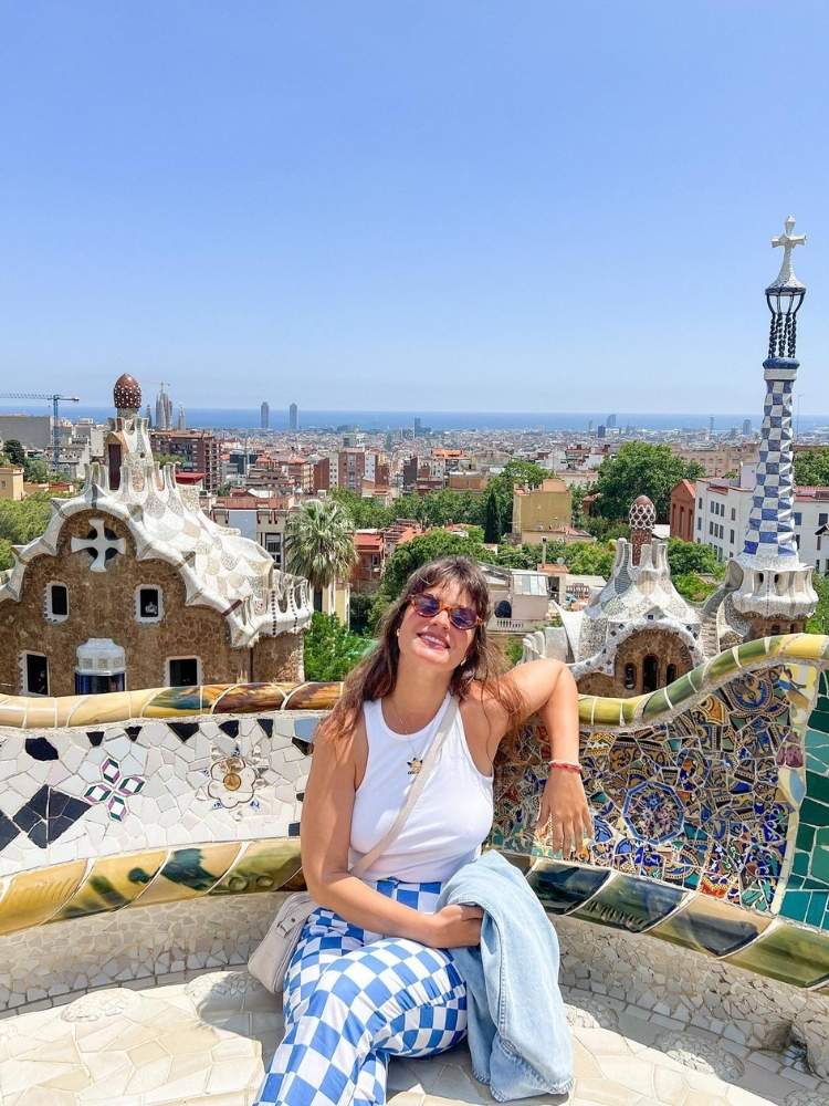 Thais Dutra Sá de regata branca, calça azul e branca, sentada no parque Gaudi com Barcelona ao fundo 