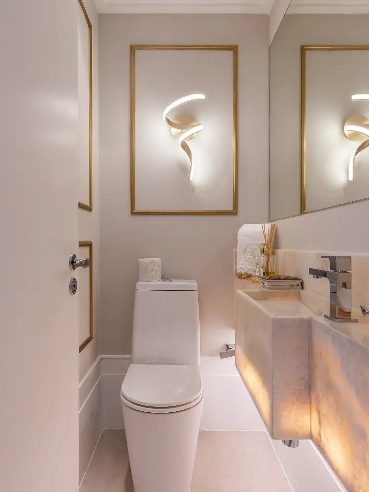 Banheiro branco com boiserie dourado, lustre, vaso sanitário, pia clara e espelho 